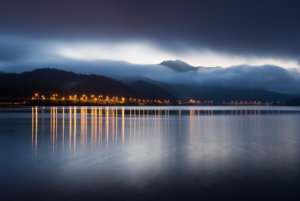 John Steele clouds roll over mountains as street lights are reflected over water